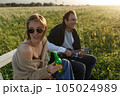 Summer time. Beautiful young couple enjoying time in field around grass. Lifestyle, love, dating, vacation concept. Woman drinking beer, man playing ukulele 105024989