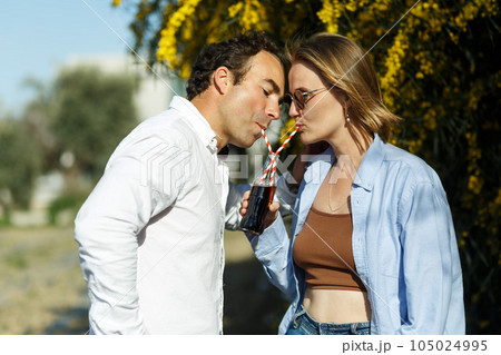 Young happy couple outdoors drinking cold drink from glass bottle with straw. Side view of man and woman drinking soft drink and smiling. Young happy couple outdoors drinking cold drink from glass bottle with straw. Side view of man and woman drinking soft drink and smiling. 105024995