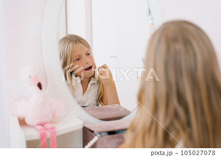 Pretty child girl paints her lips with glitter or balm in front of a mirror in her room 105027078