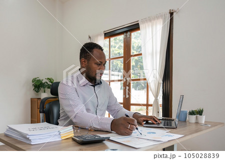 Businessman black man using laptop at home in living room. Happy mature businessman send email and working at home. African american man typing on computer with paperworks and documents on table Businessman black man using laptop at home in living room. Happy mature businessman send email and working at home. African american man typing on computer with paperworks and documents on table 105028839