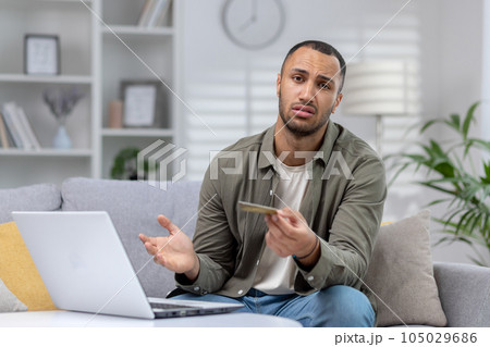Portrait of upset young African American man looking worriedly at camera and holding credit card. Sitting at home on the sofa in front of the laptop and looking disappointedly at the camera. Portrait of upset young African American man looking worriedly at camera and holding credit card. Sitting at home on the sofa in front of the laptop and looking disappointedly at the camera. 105029686