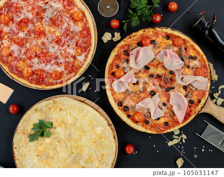 Three different pizzas on a black background with cooking ingredients. Top view, pizzeria. 105031147