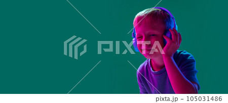 Child, little boy listening to music in headphones against cyan studio background in neon light. Grimacing face 105031486