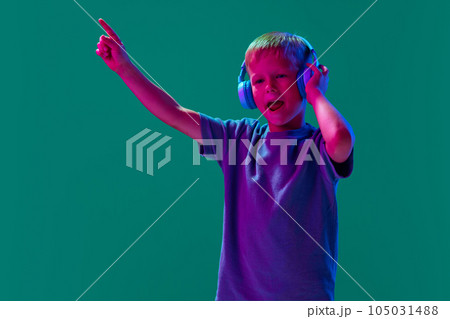Enjoyment. Child, little boy listening to music in headphones against cyan studio background in neon light 105031488