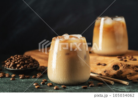 Ice coffee in a tall glass with cream poured over, ice cubes and beans on a dark concrete table. Ice coffee in a tall glass with cream poured over, ice cubes and beans on a dark concrete table. 105034062