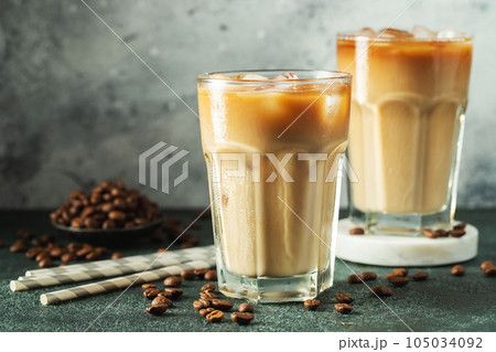 Ice coffee in a tall glass with cream poured over, ice cubes and beans on a dark concrete table. Ice coffee in a tall glass with cream poured over, ice cubes and beans on a dark concrete table. 105034092