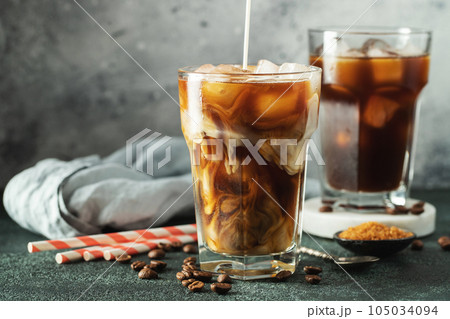 Ice coffee in a tall glass with cream poured over, ice cubes and beans on a dark concrete table. 105034094