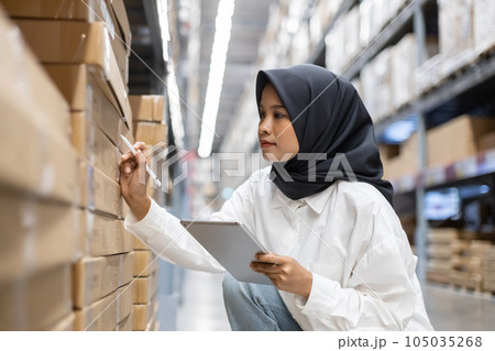 Asian muslim female headscarf hijab using digital tablet checklist product on shelves in warehouse. 105035268