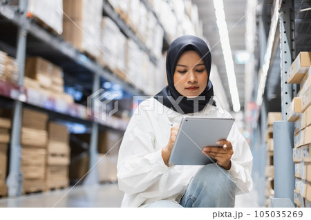 Asian muslim female headscarf hijab using digital tablet checklist product on shelves in warehouse.  105035269