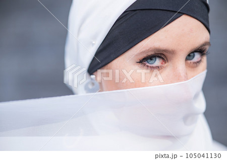 Portrait of a young blue-eyed woman in a hijab against a gray brick wall. Muslim woman with white scarf covering her face.  105041130