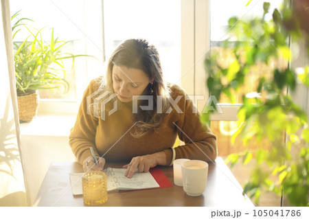 Middle-aged female poet writing in notebook creating novel or poetry while sitting at table at home 105041786