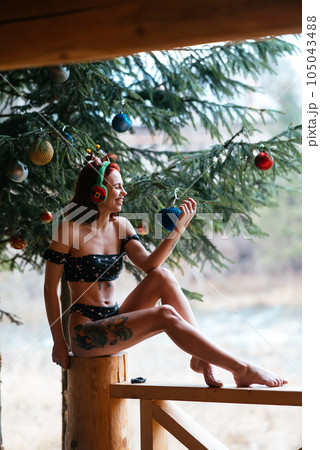 Beautiful girl in bikini sitting near Christmas tree and snow-covered trees in winter Beautiful girl in bikini sitting near Christmas tree and snow-covered trees in winter 105043488