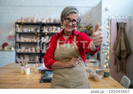 Woman potter master posing in her workshop among earthenware products showing like gesture, thumb up 105043667