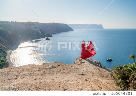 A woman in a red flying dress fluttering in the wind, against the backdrop of the sea. 105044801