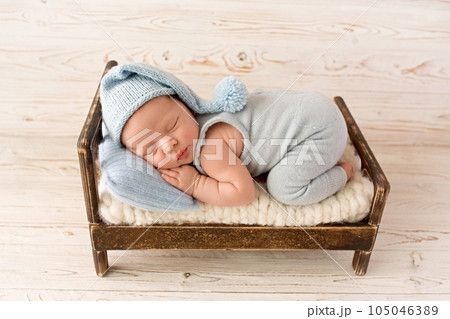 A cute newborn boy in the first days of life sleeps in a blue overalls on a wooden brown bed. Blue knitted hat and Blue head pillow. Against a light wooden background. A cute newborn boy in the first days of life sleeps in a blue overalls on a wooden brown bed. Blue knitted hat and Blue head pillow. Against a light wooden background. 105046389