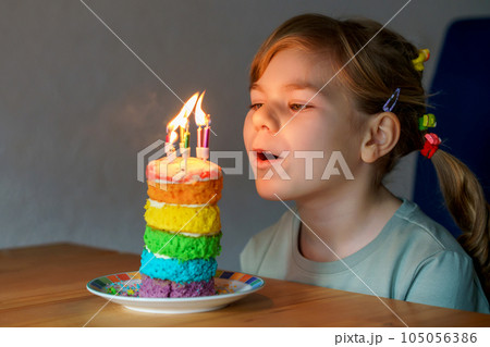 Happy little preschool girl celebrating birthday. Closeup of child with homemade rainbow cake, indoor. Happy healthy toddler blowing six candles on cake. Selective focus on cake 105056386