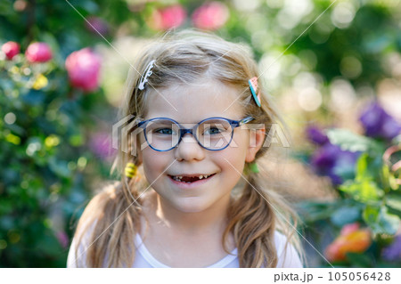 Portrait of a cute preschool girl with eye glasses and big teeth gap outdoors in park. Happy funny child on sunny summer day. Kid loosing milk tooth Portrait of a cute preschool girl with eye glasses and big teeth gap outdoors in park. Happy funny child on sunny summer day. Kid loosing milk tooth 105056428