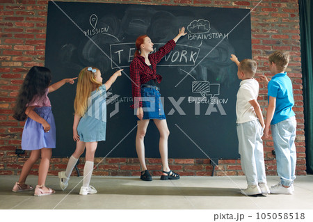 Young woman in casual clothes teaching kids to work together. Teamwork strategy icons on blackboard. 105058518