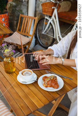 In a summer cafe, a woman surfs the net on a laptop while eating a croissant 105059427