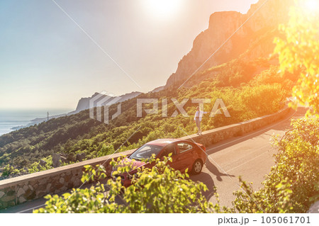 Mountain road woman. She walks along a parapet road in the middle of a mountainous area, a car is parked nearby. She is dressed in jeans and a white shirt, her hair is braided. Mountain road woman. She walks along a parapet road in the middle of a mountainous area, a car is parked nearby. She is dressed in jeans and a white shirt, her hair is braided. 105061701