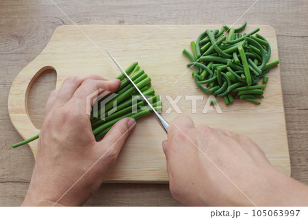 Close-up hands cutting garlic arrows for cooking and slices on a wooden board. Ingredient for Far Eastern cuisine. Top view. Close-up hands cutting garlic arrows for cooking and slices on a wooden board. Ingredient for Far Eastern cuisine. Top view. 105063997