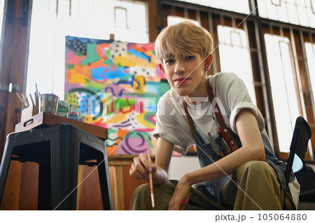 Portrait of young gay man wearing apron holding paintbrush and smiling at camera. Art, education and creative hobby concept 105064880