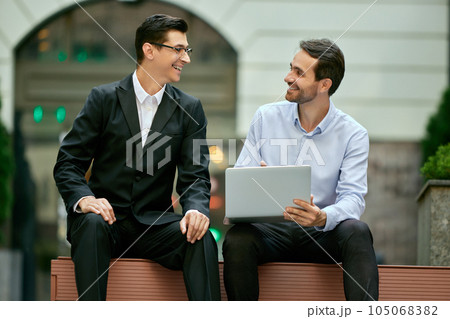 Cheerful discussion. Two businessmen, coworkers sitting on bench outside the office, looking on laptop and working 105068382