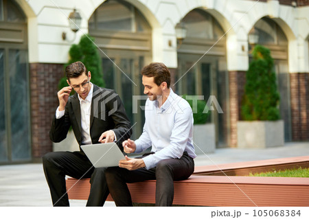 Two businessmen, coworkers sitting on bench outside the office, looking on laptop and working. Analyitcs Two businessmen, coworkers sitting on bench outside the office, looking on laptop and working. Analyitcs 105068384