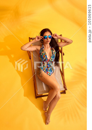 Beautiful young brunette woman in colored swimsuit lies on the lounger on yellow background Beautiful young brunette woman in colored swimsuit lies on the lounger on yellow background 105069615