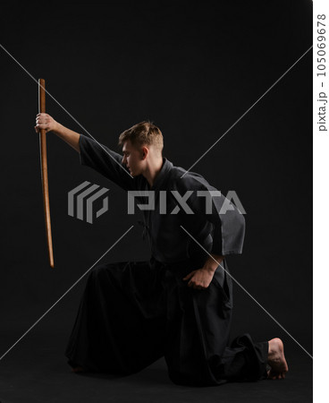 Kendo guru wearing in a traditional japanese kimono is practicing martial art with the shinai bamboo sword against a black studio background. Kendo guru wearing in a traditional japanese kimono is practicing martial art with the shinai bamboo sword against a black studio background. 105069678