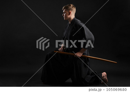 Kendo guru wearing in a traditional japanese kimono is practicing martial art with the shinai bamboo sword against a black studio background. 105069680