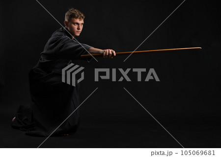Kendo guru wearing in a traditional japanese kimono is practicing martial art with the shinai bamboo sword against a black studio background. Kendo guru wearing in a traditional japanese kimono is practicing martial art with the shinai bamboo sword against a black studio background. 105069681