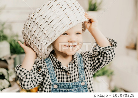 Little girl 1 years old with a basket on her head. Funny child plays puts a basket on her head like a hat and laughs, she is dressed in a plaid shirt and denim overalls. Funny moments with kids. 105070229