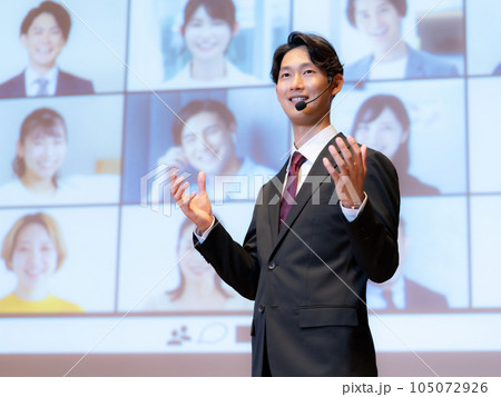 ホールで観衆とテレビ電話でつながっている人物を相手に講演をする男性講師 ホールで観衆とテレビ電話でつながっている人物を相手に講演をする男性講師 105072926