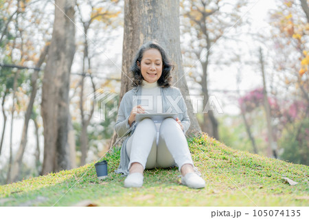 relax concepts,aged old woman Asian expressing relax, resting, stretch under tree using tablet computer and drink coffee cup in public park. relaxing relax concepts,aged old woman Asian expressing relax, resting, stretch under tree using tablet computer and drink coffee cup in public park. relaxing 105074135