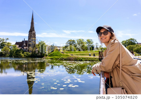 Portrait young beautiful happy smiling womane wear biege trench coat enjoy walking Kastellet citadel park against St. Alban's Church Churchill park Copenhagen Denmark . Stylish person urban lifestyle 105074820