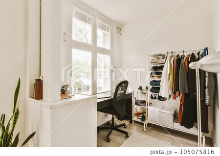 a room with a desk, chair and clothes hanging on the wall behind it is a large window that looks out onto the street 105075816
