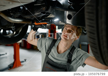 Male auto mechanic working under car lifted on elevator with flashlight 105076799