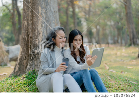 Grown daughter with aging mother expressing love and looking at tablet computer under tree in public park 105076824