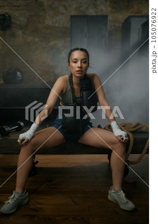 Female boxer in boxing bandage carrying gloves on shoulders sitting on bench Female boxer in boxing bandage carrying gloves on shoulders sitting on bench 105076922