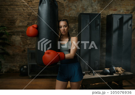 Confident young female boxer wearing gloves rest nearby punching bag Confident young female boxer wearing gloves rest nearby punching bag 105076936