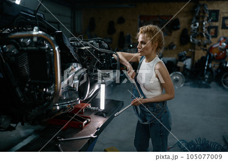 Talented girl wearing blue jumpsuit using work tool to repair motorcycle 105077009
