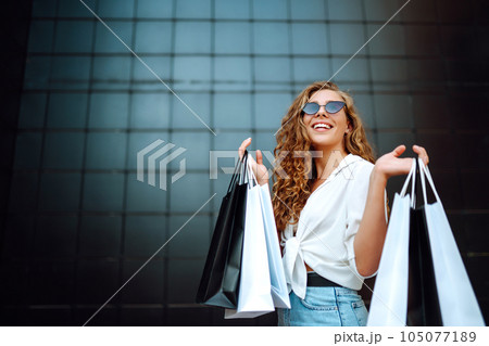 Happy stylish woman with shopping bags enjoys sunny weather walking down street. Summer shopping. 105077189
