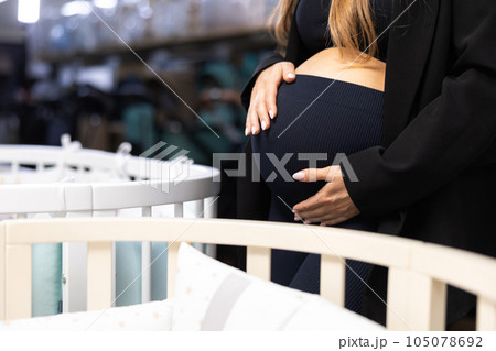 Pregnancy shopping concept. Pregnant woman choosing cradle cot or crib for newborn at baby shop store Pregnancy shopping concept. Pregnant woman choosing cradle cot or crib for newborn at baby shop store 105078692