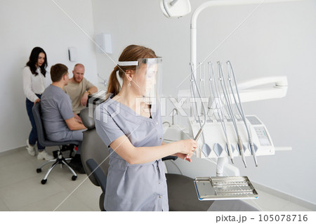 A young pretty female dentist stands in the middle of the office, and in the background a colleague is advising patients. A young pretty female dentist stands in the middle of the office, and in the background a colleague is advising patients. 105078716