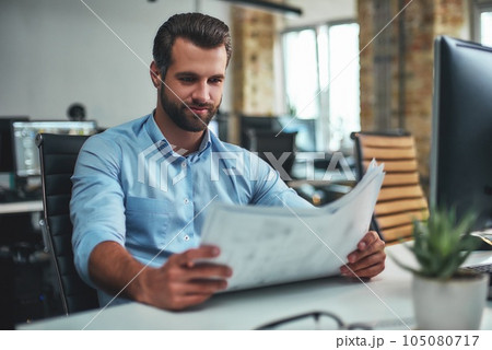 Great work Concentrated bearded engineer in formal wear looking at blueprint and smiling while sitting in the modern office 105080717
