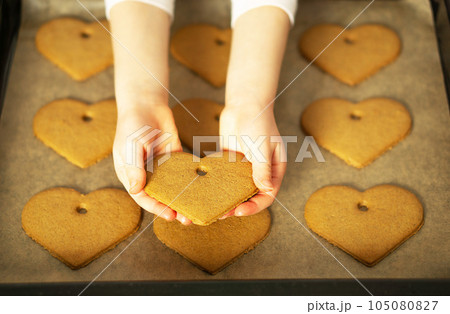 Girl hands holding a heart cookie in front of the homemade fresh baked cookies on baking sheet. Girl hands holding a heart cookie in front of the homemade fresh baked cookies on baking sheet. 105080827