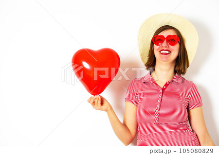 Beautiful smiling woman in red sunglasses and summer hat holding in her hand a red heart balloon. 105080829