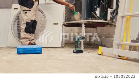 Ground level shot of trained engineer commissioned by home owner for yearly air conditioner routine maintenance, using soft dusting brush to clean dusty condenser coils and filters, outdoor Ground level shot of trained engineer commissioned by home owner for yearly air conditioner routine maintenance, using soft dusting brush to clean dusty condenser coils and filters, outdoor 105082022