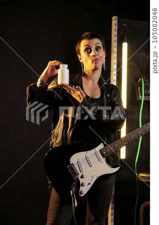 Musician holding bottle of pills pointing at health care product while recording review using modern phone, performer working at new album in sound studio. Rockstar woman playing at electric guitar 105082046
